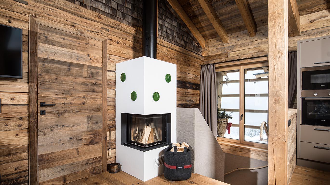 A cozy room with a modern fireplace in the center. The walls are made of wood, and large windows let in a lot of light.