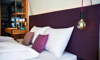 A modern bedroom with a large bed and purple cushions. An elegant lamp hangs over the headboard.
