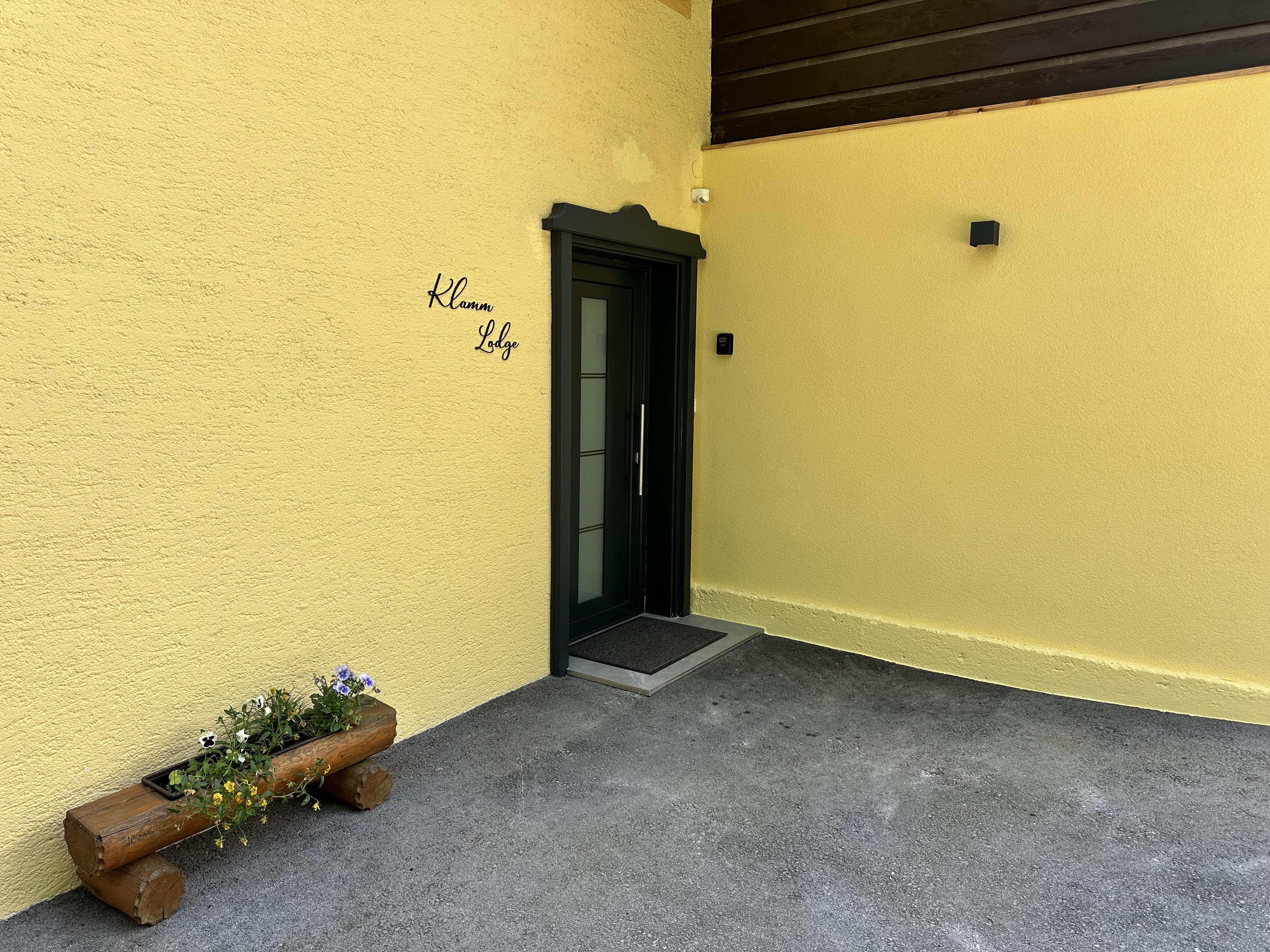 A yellow building with a black front door. On the left, there is a wooden flower box with plants.