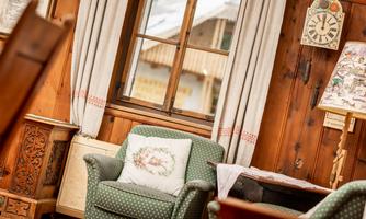 A cozy room with a green armchair and wooden paneling. Through the window, a rustic outdoor view is visible.