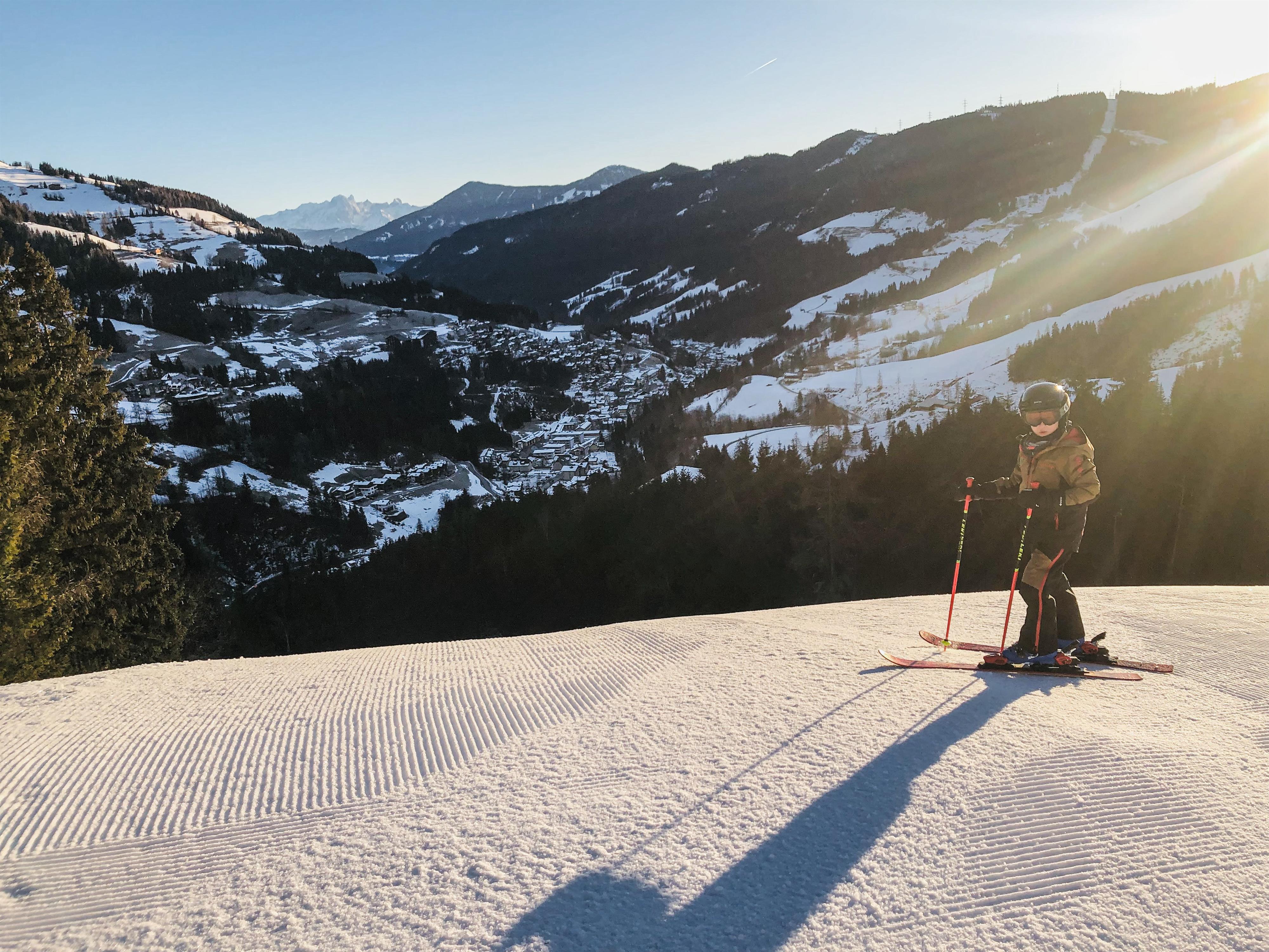 A skier stands on a snowy slope and looks down at the valley. The sun is shining over the mountains and the landscape is wintery beautiful.