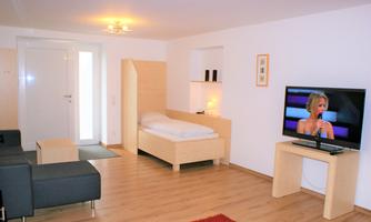 A modern living room with a single bed, a television, and a comfortable couch. The room has bright walls and a warm wooden floor.