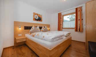 A cozy bedroom with a double bed and colorful pillows. There is a window with curtains and a colorful mural.