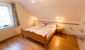 A cozy bedroom with a double bed and wooden floor. The room has windows with curtains and a small washing area.