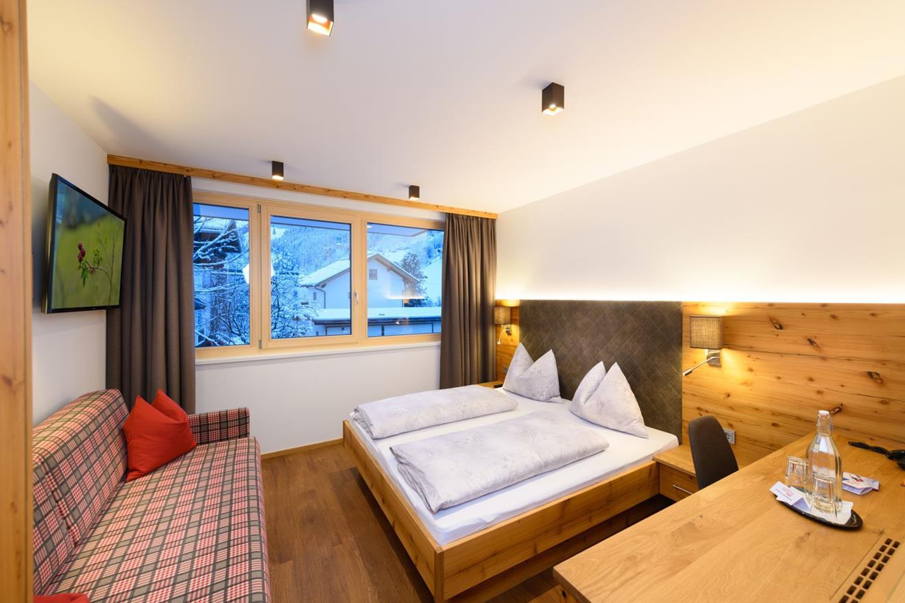 A modern hotel room with a double bed, a sofa, and a window overlooking the mountains. The warm wooden decor creates a cozy atmosphere.