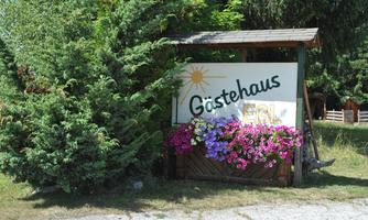 A sign reading "Guesthouse" stands in front of a beautiful flower bed. In the background, trees can be seen that create an inviting atmosphere.