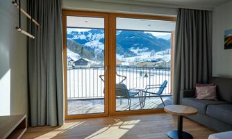 A cozy living space with large window fronts and a view of snow-covered mountains. The room is bright and modernly furnished, with a small sitting area.