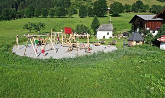 A playground with various play equipment is located in a green meadow. In the background, there are trees and a small house.