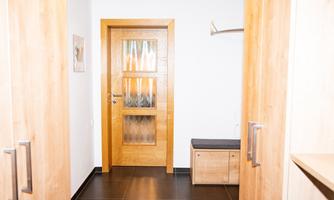 A bright hallway with wooden doors and a modern shoe cabinet. The walls are plain, and the floor is dark tiled.