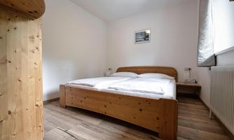 A simple bedroom with a wooden bed and a nightstand. The walls are light, and there is a window with curtains.