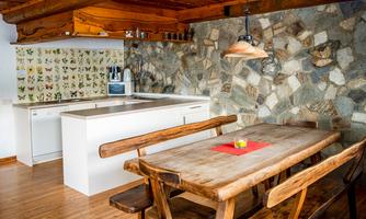 A cozy kitchen with a large wooden table and rustic chairs. The walls are adorned with natural stone and a colorful wallpaper.