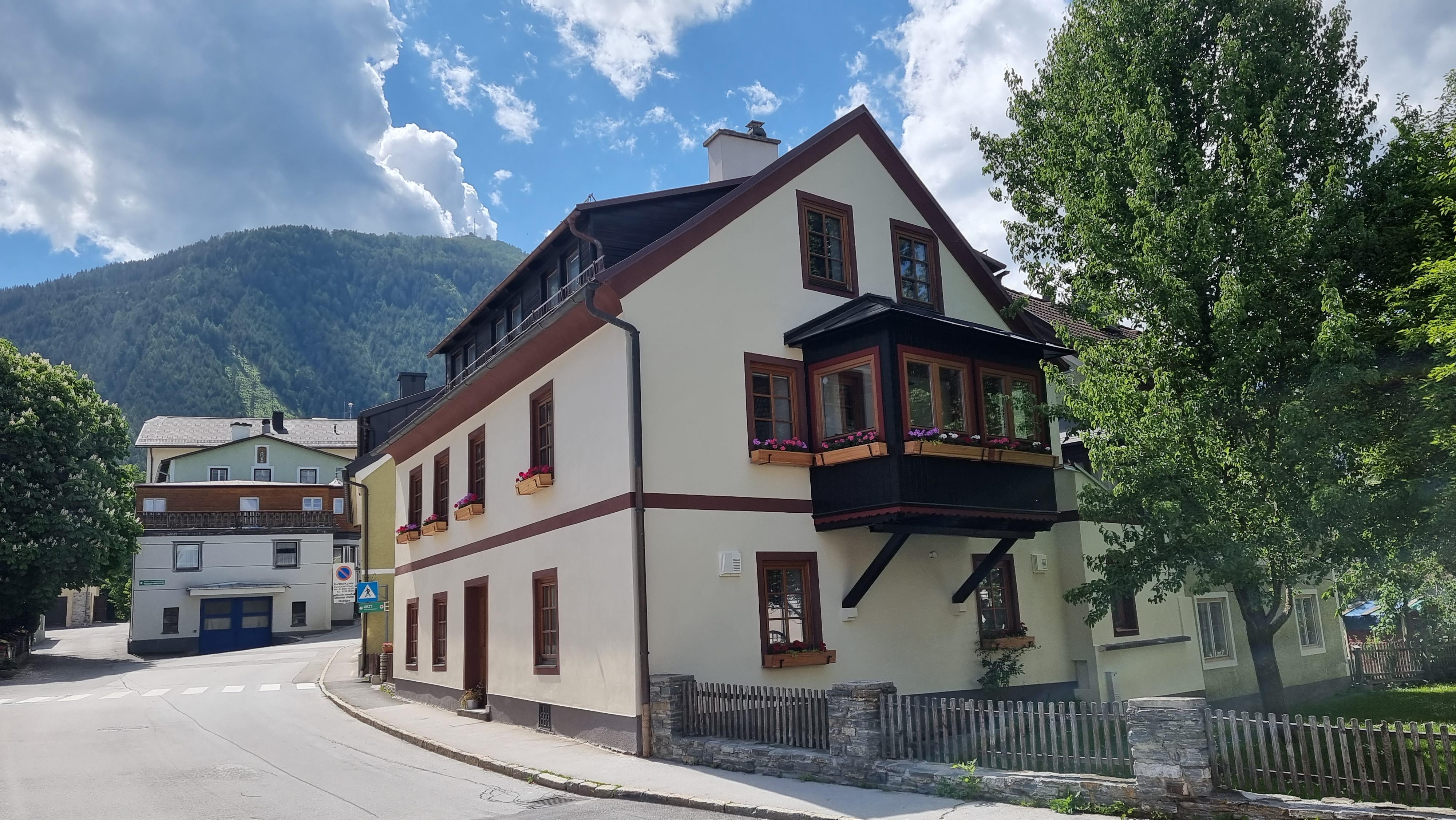 A charming house on a street corner with a balcony and beautiful windows. In the background, mountains and a blue sky are visible.