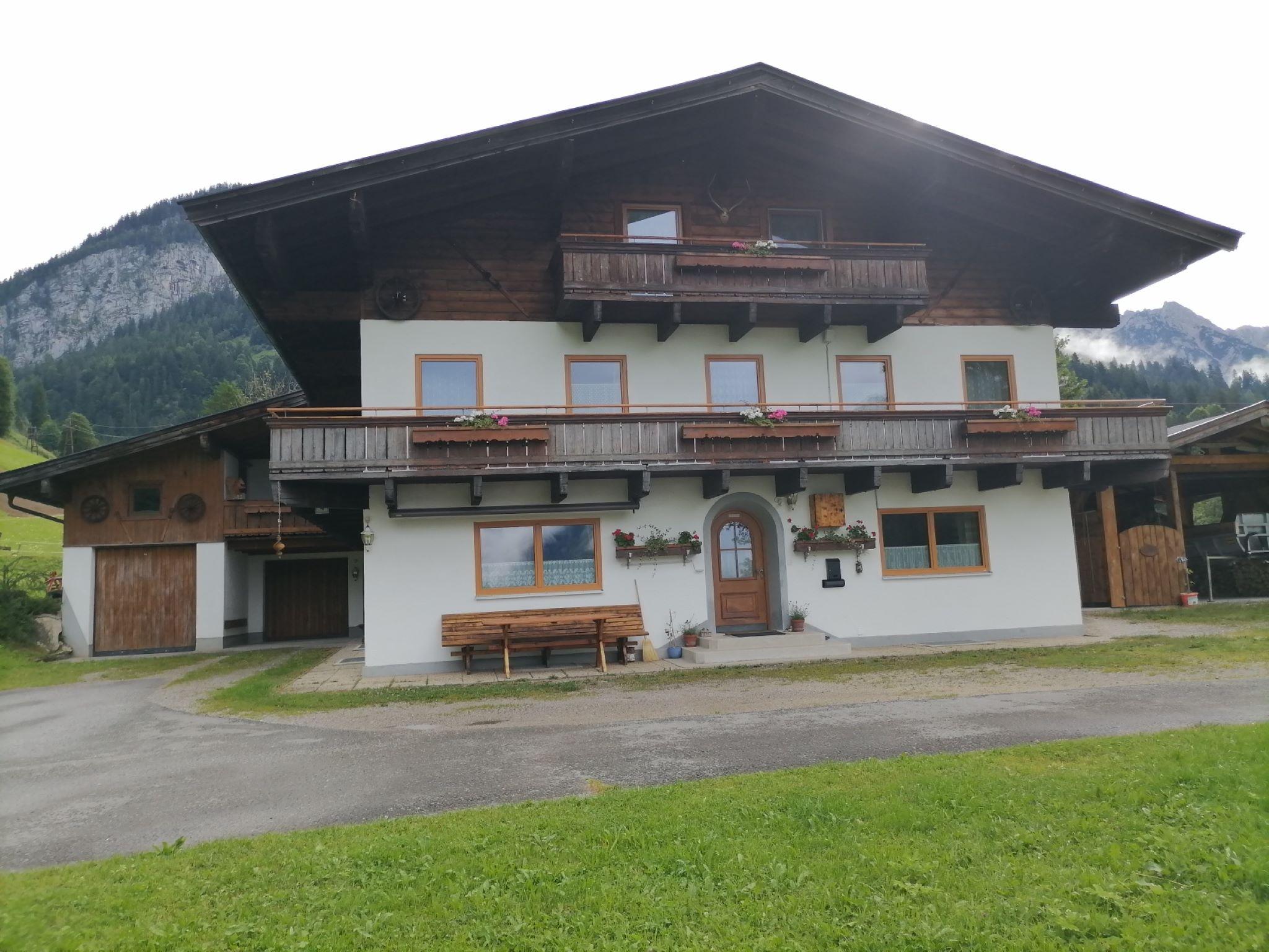 A traditional alpine house with several balconies and a large meadow in front. In the background, mountains can be seen, creating a picturesque landscape.