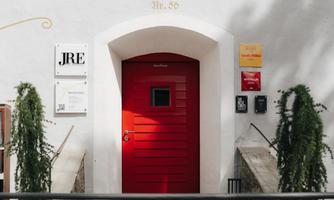 A red door in a white wall with several signs beside it. It is an inviting entrance situation.