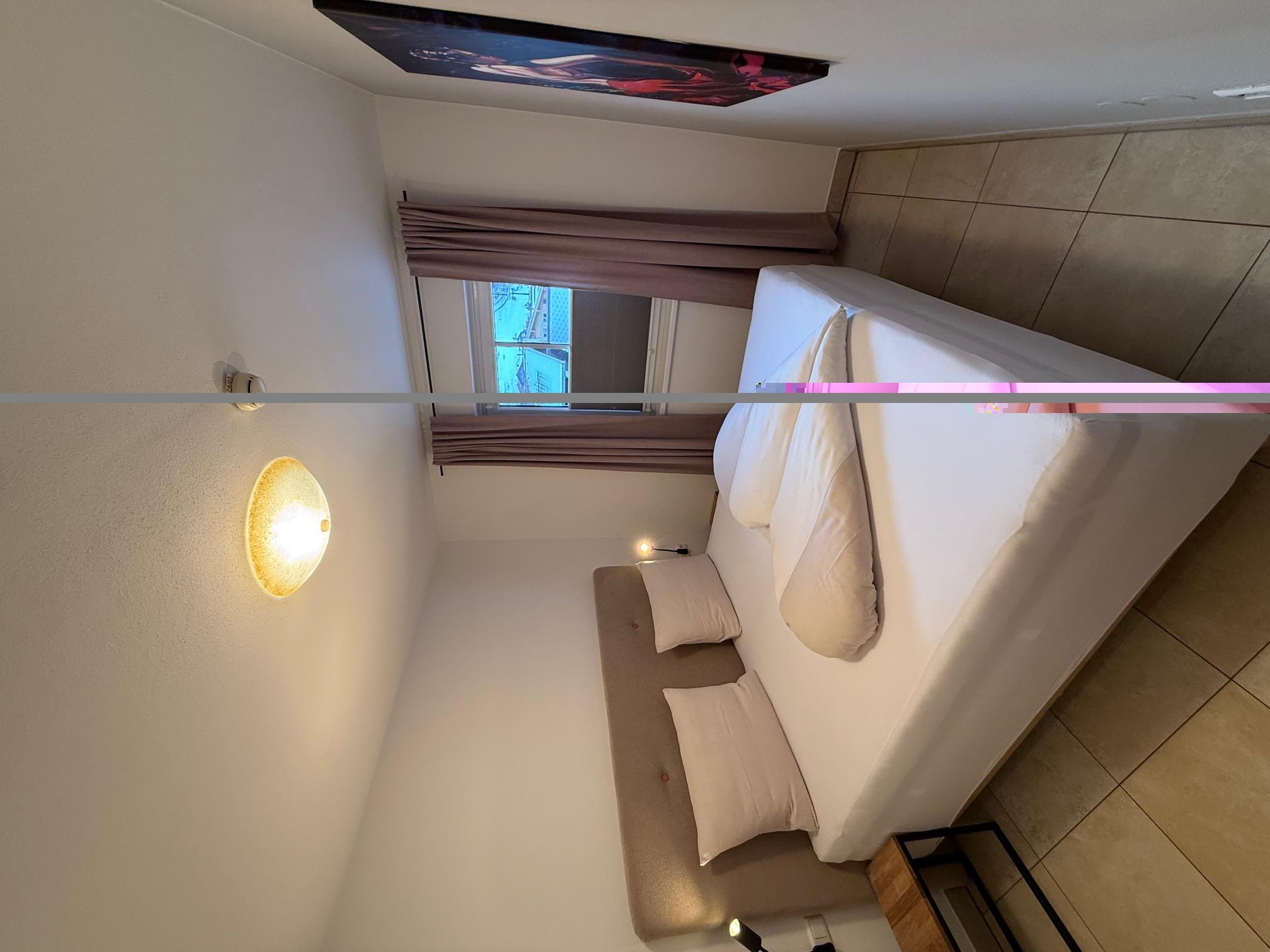 A cozy bedroom with a double bed and two pillows. The walls are light, and there is a window with curtains.