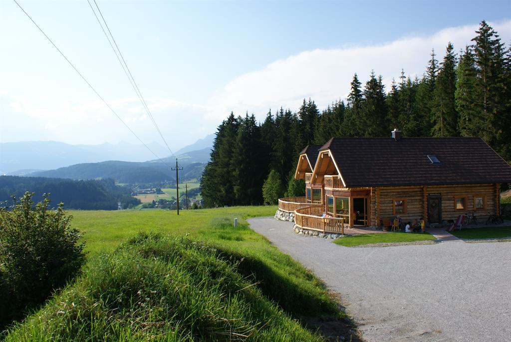 A cozy wooden house stands at the edge of a meadow, surrounded by trees and gentle hills. In the background, the mountains stretch out under a clear sky.