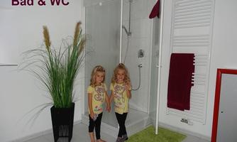 Two girls are standing in a modern bathroom next to a shower. In the background, a large plant and a towel rack are visible.