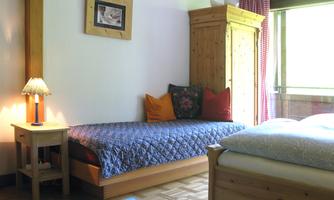 A cozy room with two beds and a wooden piece of furniture. There is a small bedside table with a lamp and a window that lets in a lot of light.