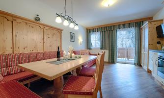 A cozy dining area with a large table and red upholstered chairs. The room is bright and friendly with wood paneling and a view of the balcony.