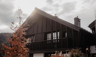 A modern, dark wooden house with a distinctive balcony cladding. In the foreground, autumnal plants and a cloudy sky can be seen.