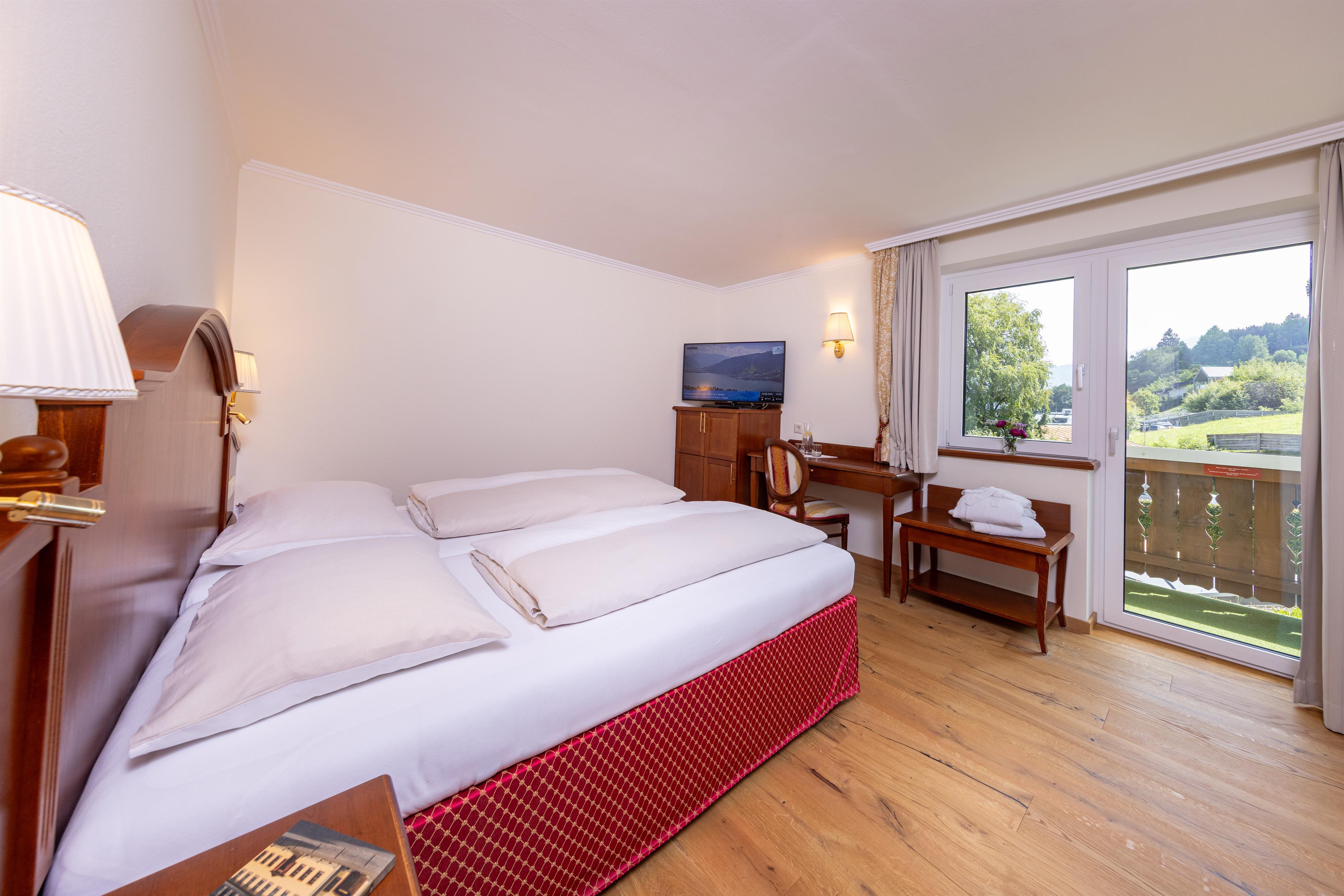 A cozy hotel room with two beds and a wooden table. Large windows offer a beautiful view of the greenery.