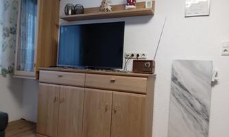 A modern living room with a large television on a wooden cabinet. A decorative element hangs on the wall and there is a windowsill with a small plant.
