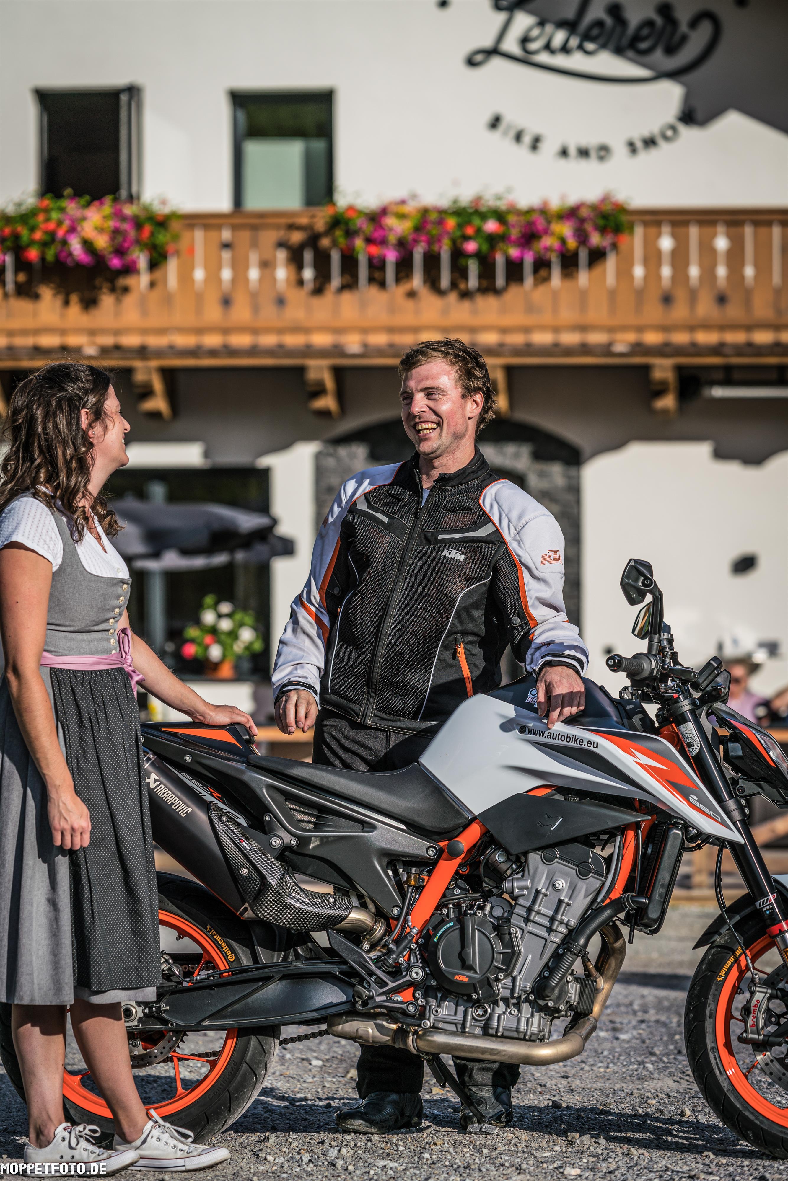 A motorcyclist in protective clothing stands next to his bike and speaks with a woman in traditional attire. Behind them, a building with flowers and a sign can be seen.