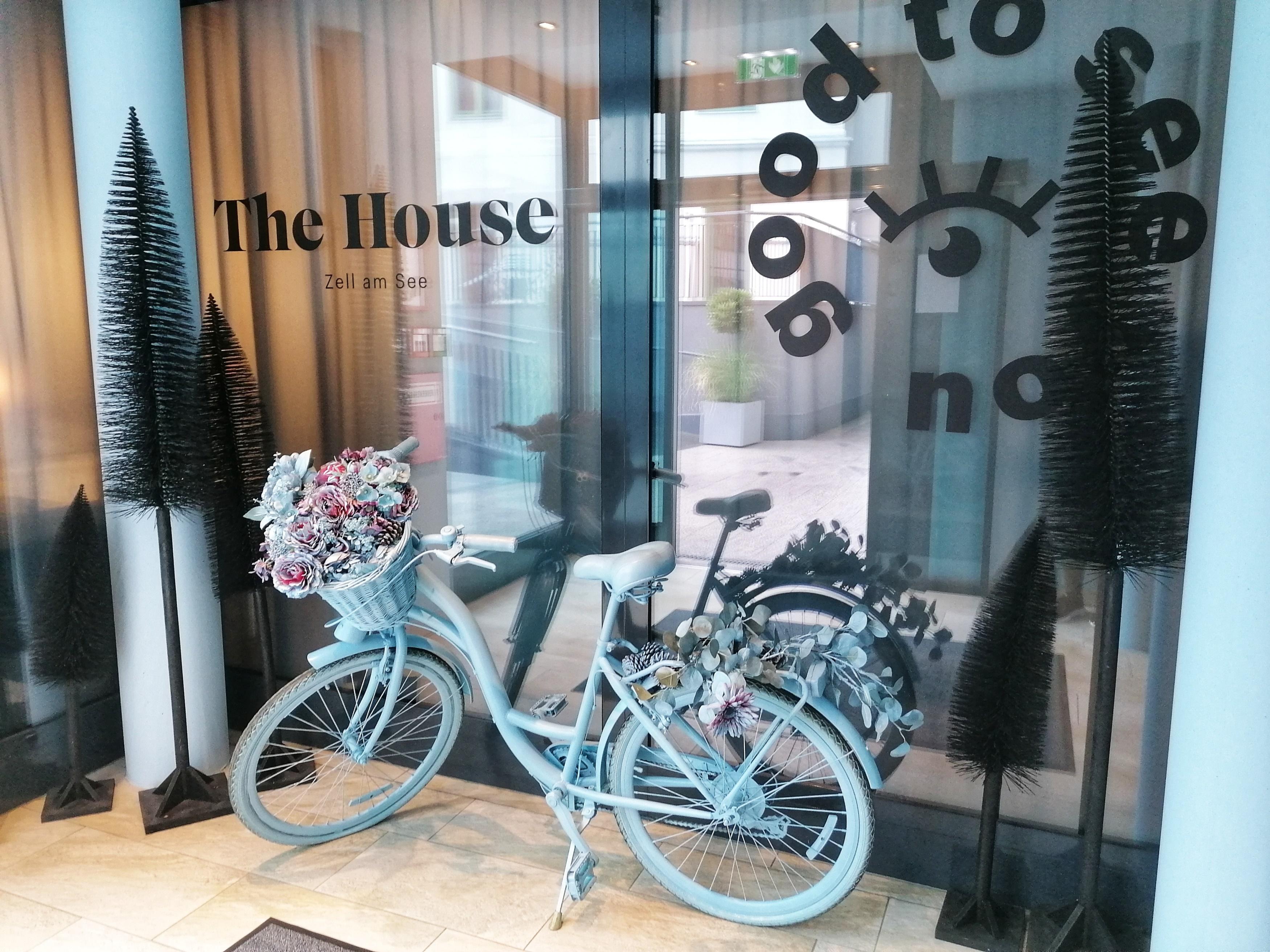 A stylish entrance with a white shower and a bicycle decorated with flowers. In the background, slender plants and windows with inscriptions can be seen.