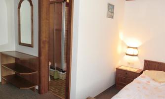 A simple bedroom with a bed, a nightstand, and a wardrobe. In the background, a bathroom with a shower can be seen.