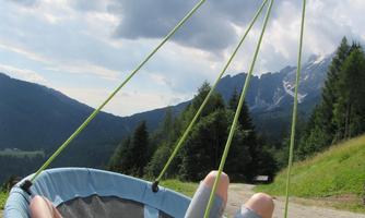 A child lies relaxed in a large hammock with a view of green hills and mountains. The sky is partially cloudy and the atmosphere feels peaceful.
