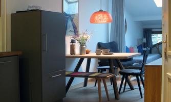 A cozy dining kitchen with a round table and modern chairs. A bright lamp hangs above the table, and the room has large windows for natural light.