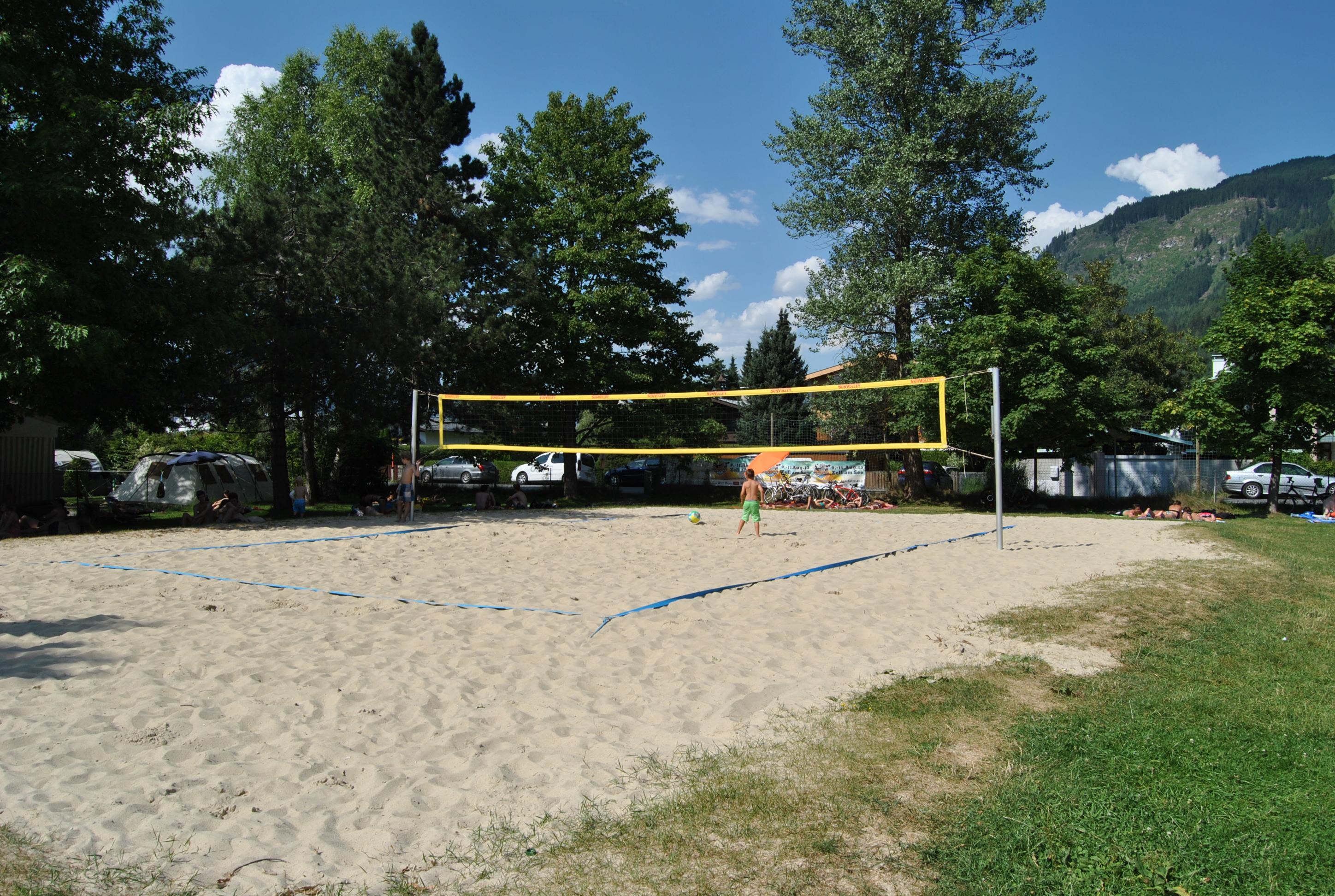 A volleyball court made of sand surrounded by trees and grass. It is sunny and clear skies are visible.