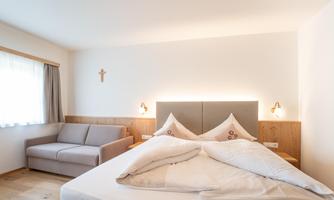 A bright bedroom with a large bed and a gray sofa. The walls are neutral, and there is a small cross decoration.