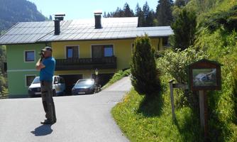 A person is standing on a path in front of a yellow house and taking pictures. In the background, trees and mountains can be seen.
