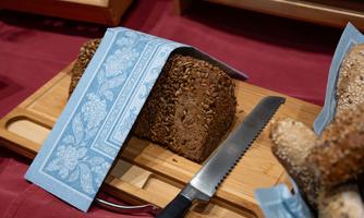 A freshly baked bread on a wooden board, with a blue cloth and a knife beside it. The bread has a rustic, grainy surface.