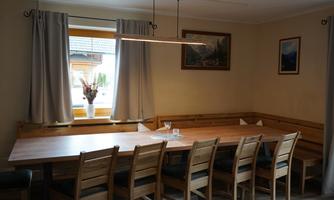 A cozy dining area with a large wooden table and numerous chairs. The windows are adorned with curtains and pictures hang on the wall.