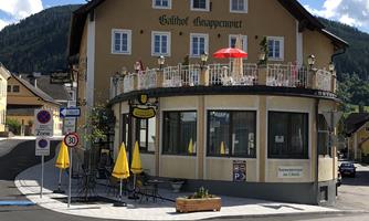 A characteristic inn at a street corner with a large balcony and sun umbrellas. In the background, green hills and a clear sky can be seen.