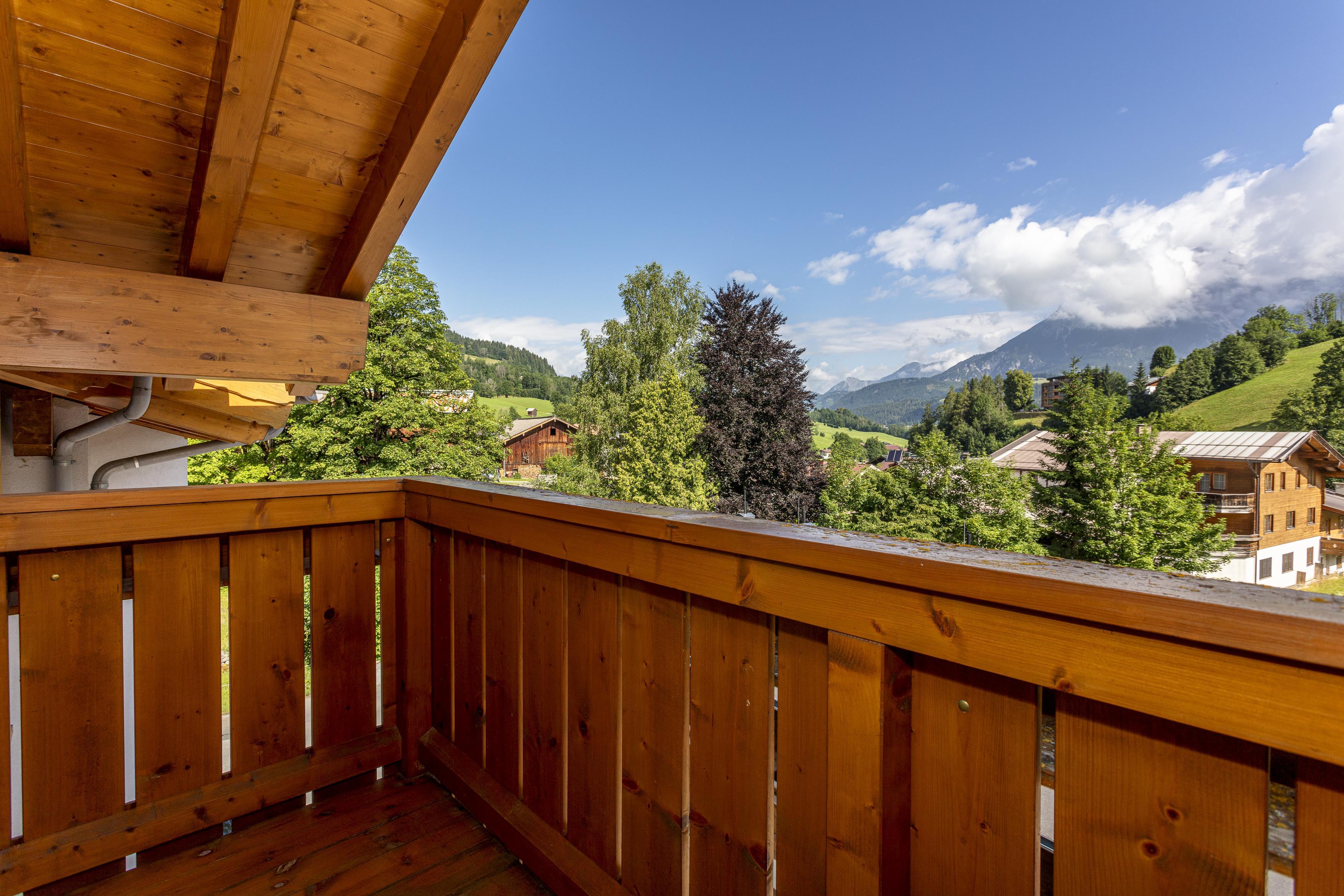 A balcony with a wooden railing and a view of a picturesque landscape. In the background are forests and mountains under a radiant blue sky.