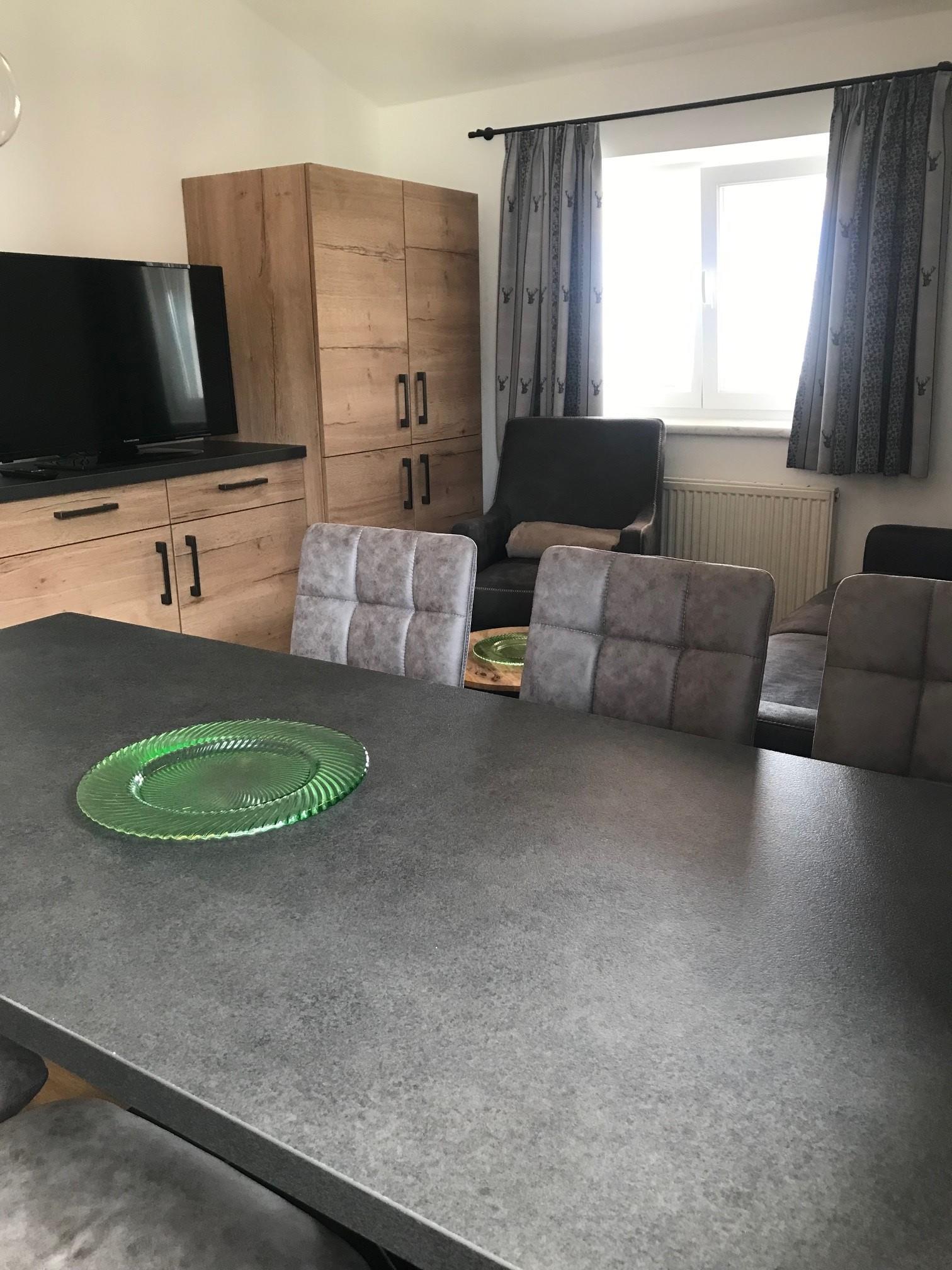 A modern living room with a large dining table and chairs. In the background, a television and a window are visible.