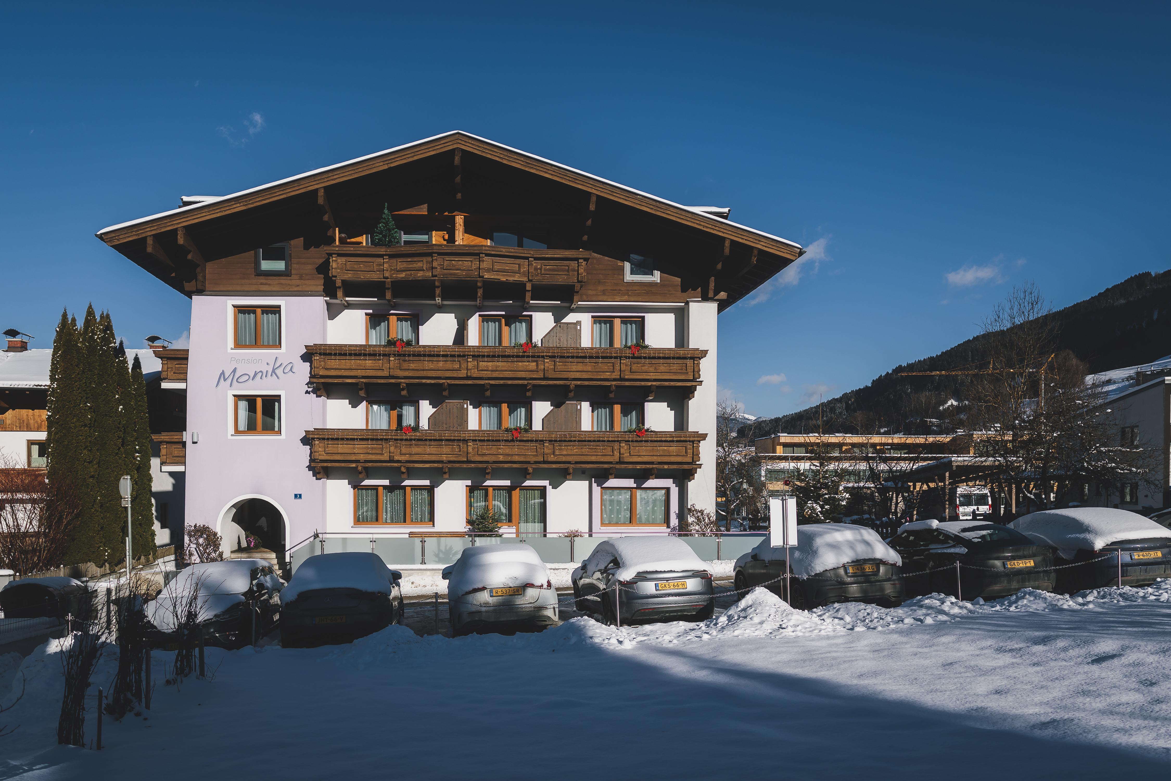 A large Alpine building with several balconies, surrounded by snow. In the foreground, cars are seen under a layer of snow.