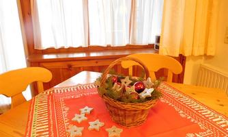 A cozy table with a red tablecloth and a basket full of Christmas decorations. The windows are adorned with bright curtains that let light shine in.
