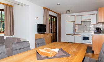 A modern kitchen with a large dining table and comfortable chairs. There is a television and patio doors that let in light.