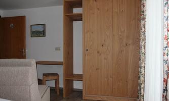 A simply furnished room with a wooden wardrobe and a small table. A beige armchair is also visible, and the walls are painted light.