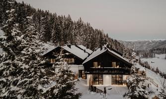 A modern house in the snow, surrounded by tall, green firs. The sky is bright and the landscape looks calm and inviting.