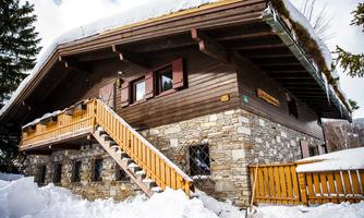 A cozy holiday home made of wood and stone, surrounded by snow. The staircase leads to the front door, and the facade is adorned with windows and a protruding roof.
