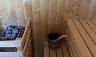A cozy sauna with wooden-paneled walls and benches. Next to the stones, there is a bucket with a ladle.