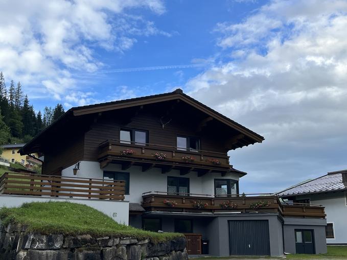 A beautiful alpine house with wooden cladding and a balcony. In the background, green hills and a blue sky can be seen.