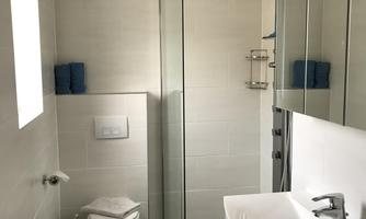 A modern bathroom with a glass shower and a sink. The walls are light and the tiles are dark, with blue towels as an accent.