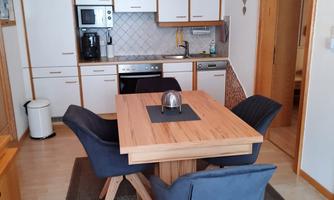 A modern kitchen with a dining area in the center. The table is made of wood, surrounded by blue chairs, and the room design is bright and friendly.