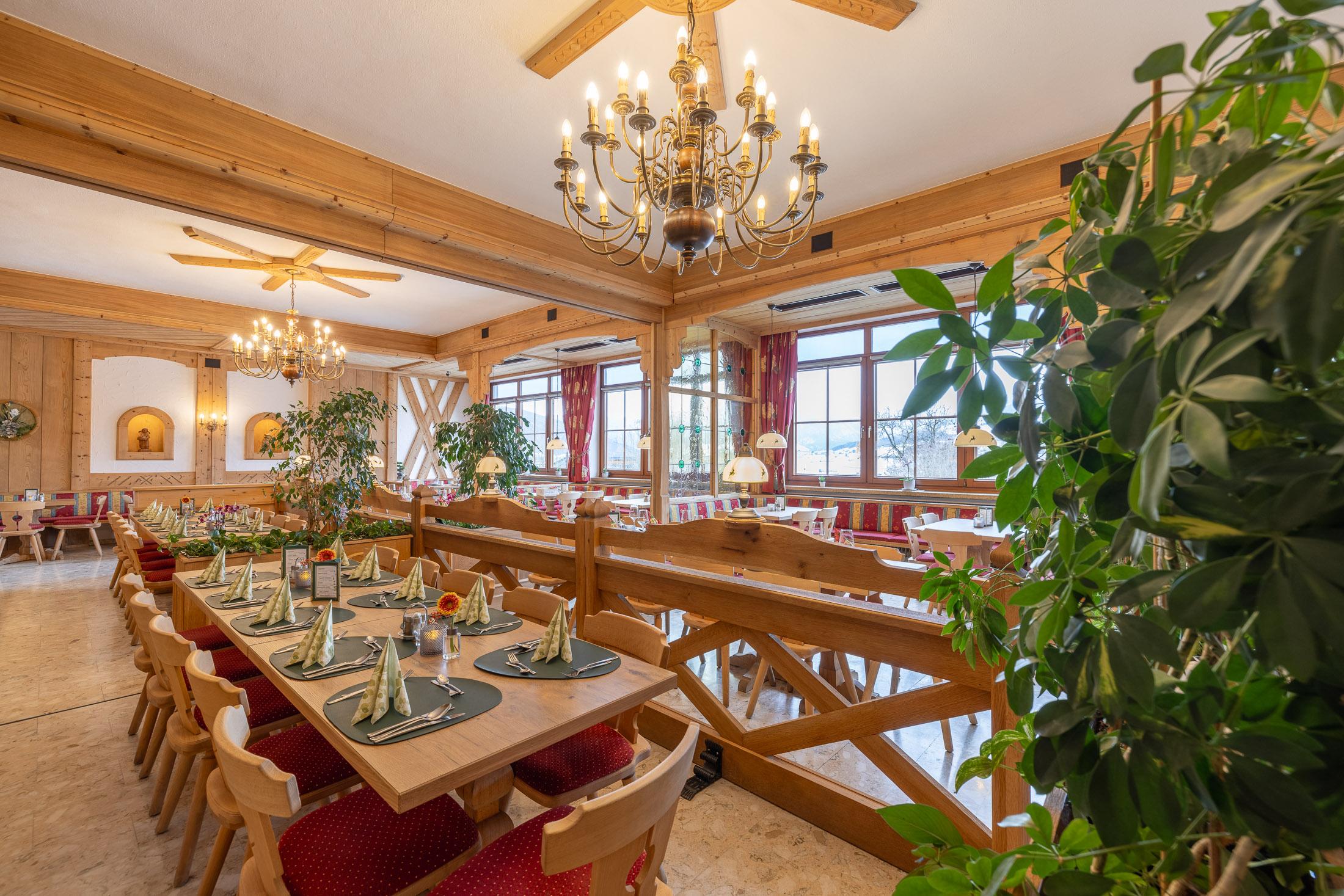 A cozy restaurant with wooden elements and large windows. The tables are elegantly set and surrounded by plants.